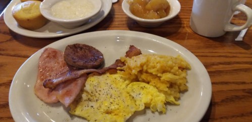 A breakfast plate with scrambled eggs, bacon, sausage, ham, and hash browns, served with grits and fruit on the side.