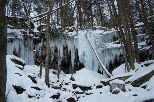 A frozen waterfall surrounded by snow-covered rocks and trees in a winter forest setting.