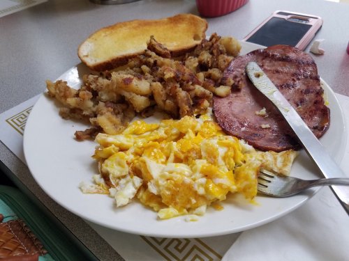 A plate of breakfast featuring scrambled eggs, hash browns, ham, and a slice of toast.