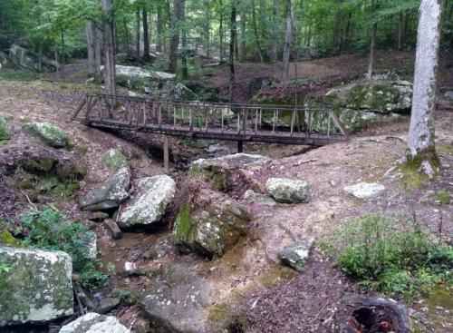 A wooden bridge spans a small creek in a forested area, surrounded by rocks and greenery.