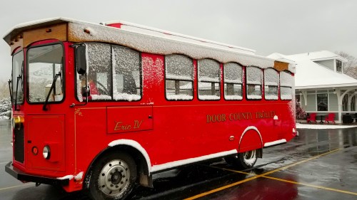A red trolley bus covered in snow parked outside a building on a cloudy day.