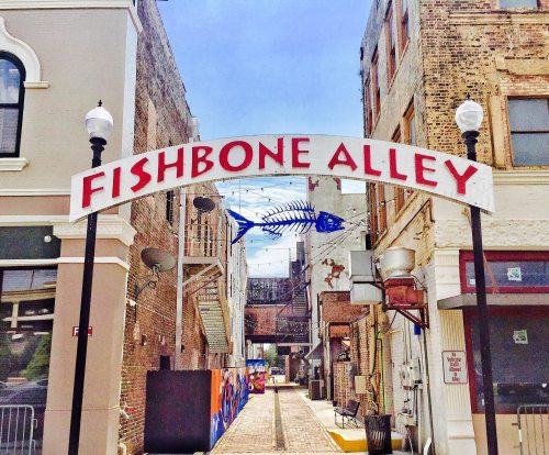Sign for Fishbone Alley, a narrow street with brick pavement and colorful murals, flanked by buildings.