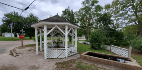 A white gazebo with a small water feature, surrounded by greenery and a stop sign in a quiet neighborhood.