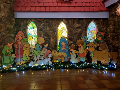 Colorful cartoon figures of children and animals gathered around a nativity scene, with stained glass windows in the background.
