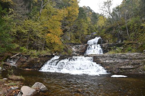 A serene waterfall cascades over rocky terrain, surrounded by autumn foliage and a calm river below.