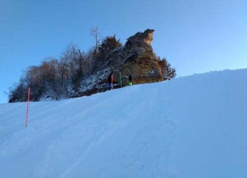 Two skiers near a rocky outcrop on a snowy slope, with clear blue skies above.