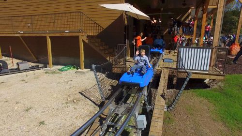 A child rides a blue roller coaster on a track near a wooden building, with people gathered around.