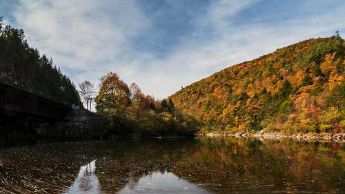 A serene river reflects colorful autumn foliage and a hillside under a clear blue sky.