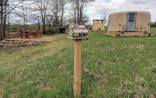 A wooden birdhouse on a post in a grassy area, with a yurt and outdoor seating in the background.
