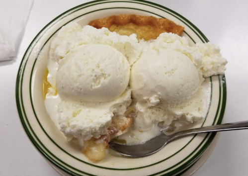 A bowl of dessert featuring two scoops of vanilla ice cream atop a warm fruit pie. A spoon rests beside the bowl.