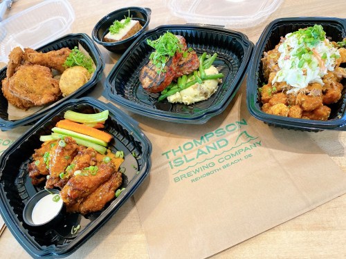 Four takeout containers of food featuring fried chicken, grilled meat, green beans, and noodles, with a logo from Thompson Island Brewing Company.