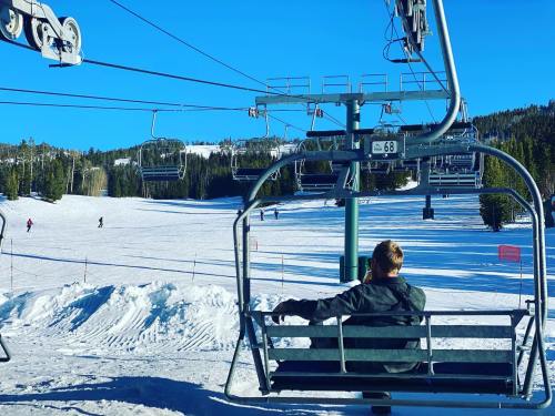 A person sits on a ski lift, overlooking a snowy slope with skiers in the distance and trees under a clear blue sky.