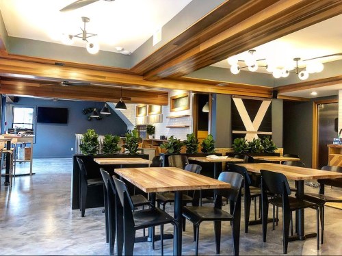 Modern dining area with wooden tables, black chairs, and greenery, featuring stylish lighting and a sleek kitchen backdrop.