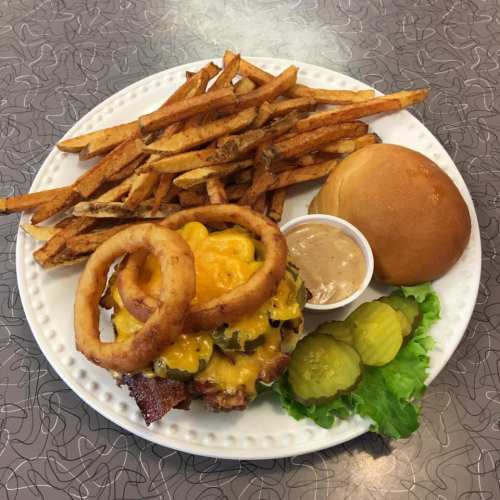 A plate with a cheeseburger topped with onion rings, pickles, lettuce, a side of fries, and a bun with dipping sauce.