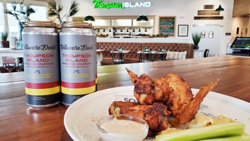 A plate of chicken wings with celery and ranch dressing, alongside two cans of Thompson Island Brewing Company beer.