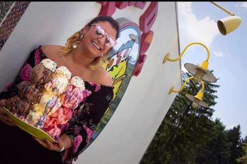 A smiling woman holds a large platter of colorful ice cream scoops, wearing sunglasses and a black off-shoulder top.