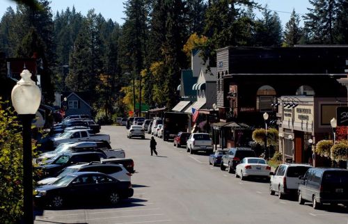 A quiet street lined with shops and parked cars, surrounded by trees under a clear blue sky.