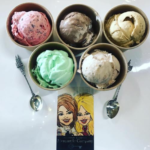 Five cups of colorful ice cream in various flavors, with two spoons and a cartoon card featuring two women.