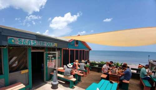 Outdoor dining area at a beachside restaurant with colorful tables, patrons enjoying meals, and ocean views.