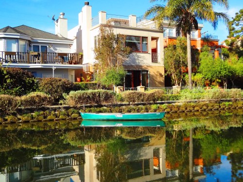 A green kayak floats on a calm canal, reflecting colorful houses and palm trees under a clear blue sky.