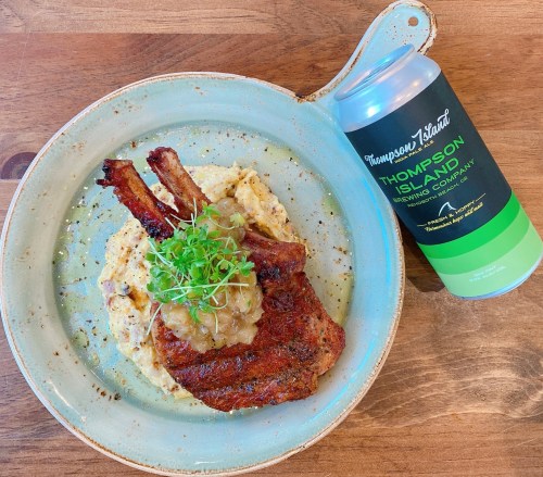 A plate with grilled pork chop on creamy mashed potatoes, topped with sauce and microgreens, alongside a can of Thompson Island beer.