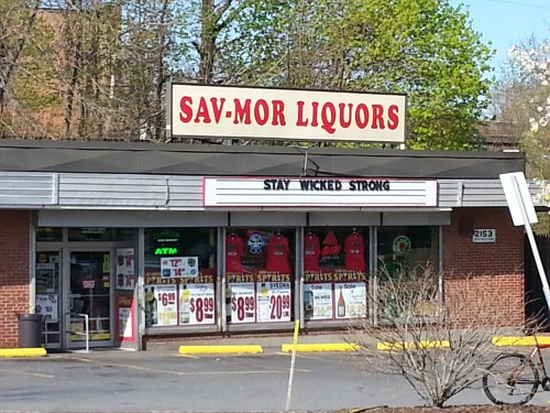 Exterior of Sav-Mor Liquors store with a sign reading "STAY WICKED STRONG" and various promotional banners.
