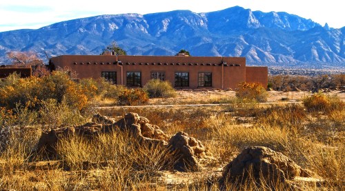 A rustic adobe building surrounded by dry grass and rocky formations, with mountains in the background under a clear sky.