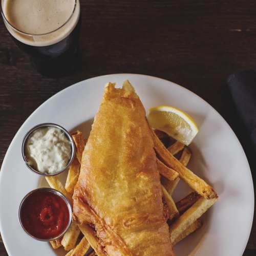 A plate of golden-battered fish and fries, served with tartar sauce, ketchup, and a lemon wedge, alongside a dark beer.