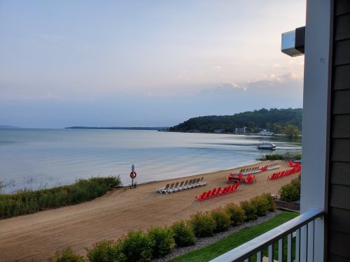 A serene lakeside view with red chairs lined up on the sandy shore and boats in the distance under a soft sky.