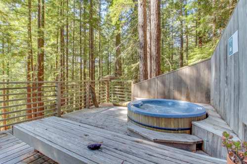 A wooden deck with a hot tub surrounded by tall redwood trees in a serene forest setting.