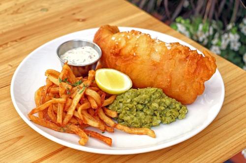 A plate of crispy fish with fries, mushy peas, a lemon wedge, and a small cup of dipping sauce on a wooden table.