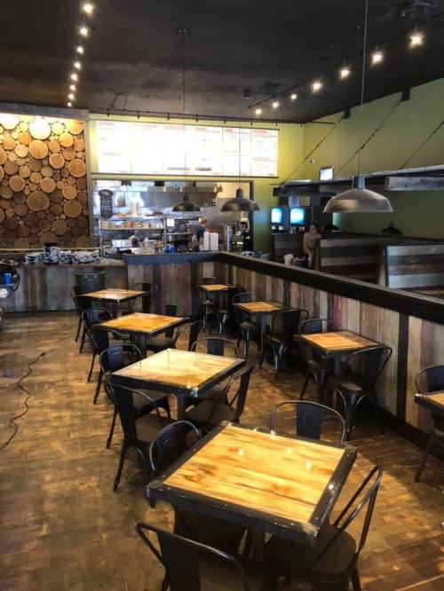 Interior of a restaurant featuring wooden decor, metal chairs, and tables, with a visible kitchen area in the background.