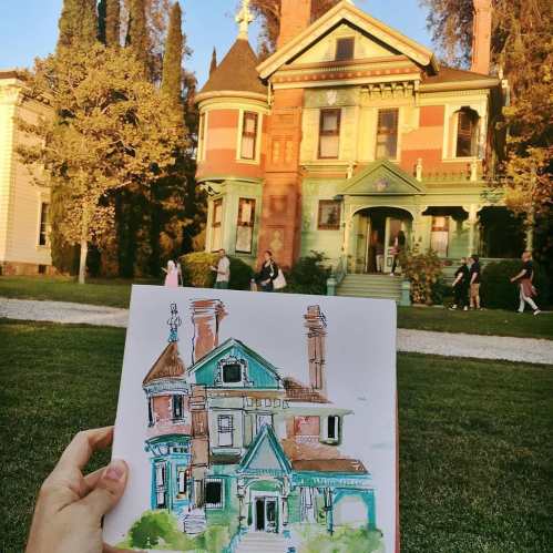 A hand holds a colorful sketch of a Victorian house in front of the actual house, with people walking in the background.