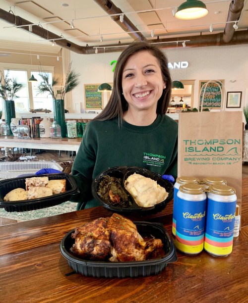 A smiling woman holds takeout containers of food and drinks from Thompson Island Brewing Company in a cozy setting.