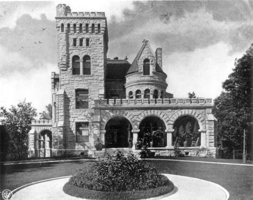 Historic stone castle with a tower, arched entrance, and surrounding trees, set against a cloudy sky.