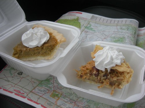 Two slices of pie topped with whipped cream, resting on a map in a car. One is a green pie, the other a coconut pie.