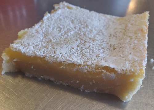 A square piece of lemon bar dessert dusted with powdered sugar on a metallic surface.