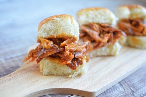 Three pulled pork sliders on a wooden board, featuring tender meat in a soft, golden bun.