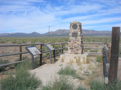 A stone monument surrounded by a fence, with informational signs, set against a backdrop of mountains and blue sky.
