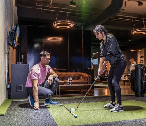 A golf instructor watches a student practice putting in a modern indoor facility.