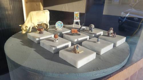 A display featuring various small figurines and objects on white pedestals, including a cow and a map.