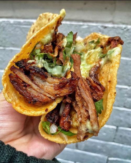 A hand holding a taco filled with grilled meat, cilantro, onions, and a drizzle of green sauce against a brick background.