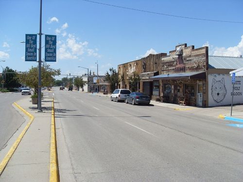 A quiet street scene featuring a saloon and shops, with blue skies and a few parked cars.