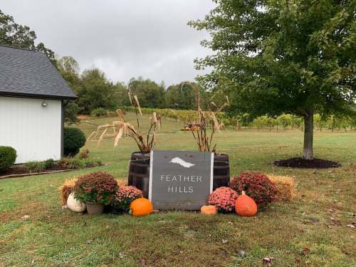 Sign for "Feather Hills" surrounded by pumpkins, flowers, and decorative corn, set in a grassy area with trees in the background.