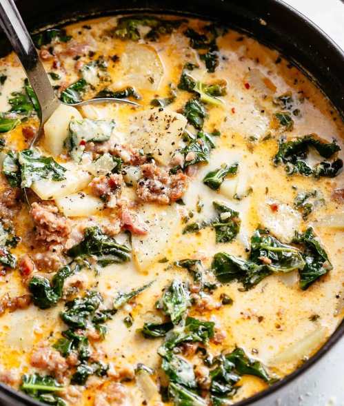 A creamy soup with potatoes, sausage, and leafy greens, garnished with black pepper in a dark pot.