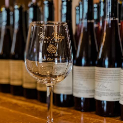 A clear wine glass with "Cave Ridge Vineyard" etched on it, set against a backdrop of wine bottles.