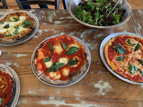 Two pizzas topped with basil and cheese, alongside a bowl of mixed greens on a rustic wooden table.