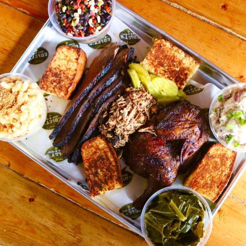 A wooden table with a platter of barbecue meats, sides, and cornbread, including pulled pork, brisket, and collard greens.