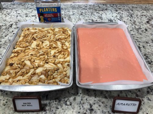 Two trays of candy: one with salted nut roll topped with peanuts, the other with pink amaretto fudge. Planters peanuts in background.