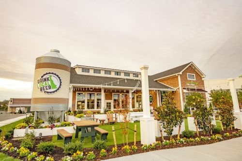 A modern brewery with a large beer silo, outdoor seating, and landscaped gardens under a cloudy sky.